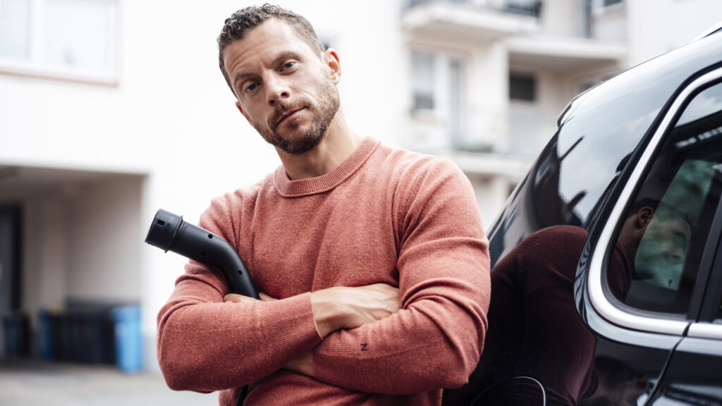 Bister man med armarna i kors står intill en bil med en laddsladd.