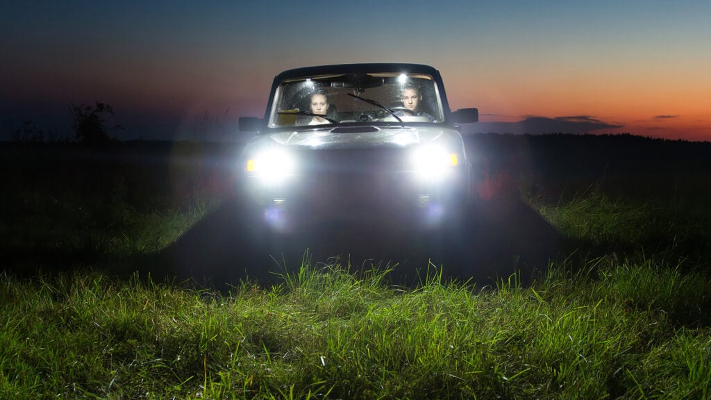 Två personer i en äldre bil med tända strålkastare.
