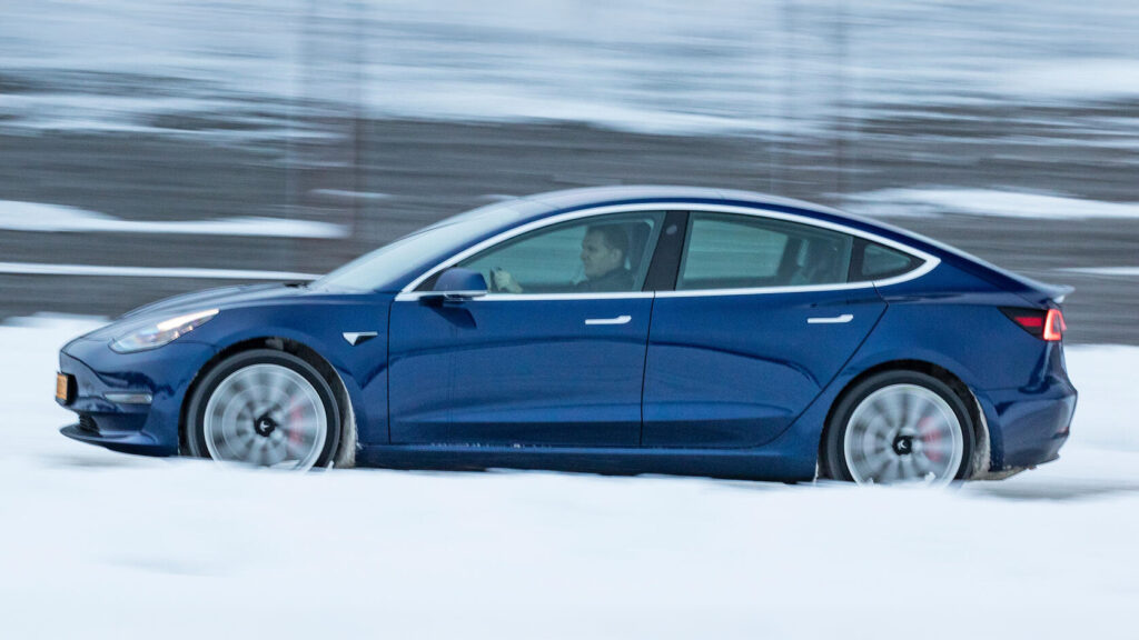 Blå Tesla Model 3 i profil i snöig omgivning.