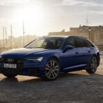 Blå Audi A6 Avant med hamn och solnedgång i bakgrunden.