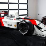 En livs levande replika av Ayrton Sennas McLaren MP4/4 från 1988, placerad på en svart duk i en garage-miljö. Modellen är i full storlek med detaljerad livery, inklusive Marlboro, Shell och Goodyear-logotyper.