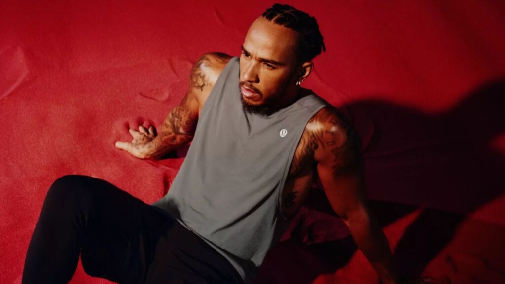 Lewis Hamilton sitter på en röd matta i träningskläder från Lululemon, iförd en grå ärmlös tröja och svarta tights.