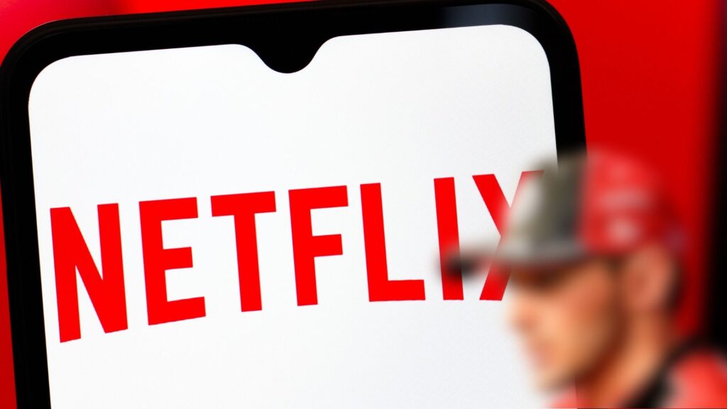 En stor Netflix-logga på en mobilskärm, med en suddig F1-förare i röd keps i förgrunden.