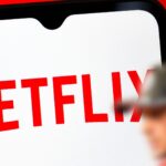 En stor Netflix-logga på en mobilskärm, med en suddig F1-förare i röd keps i förgrunden.