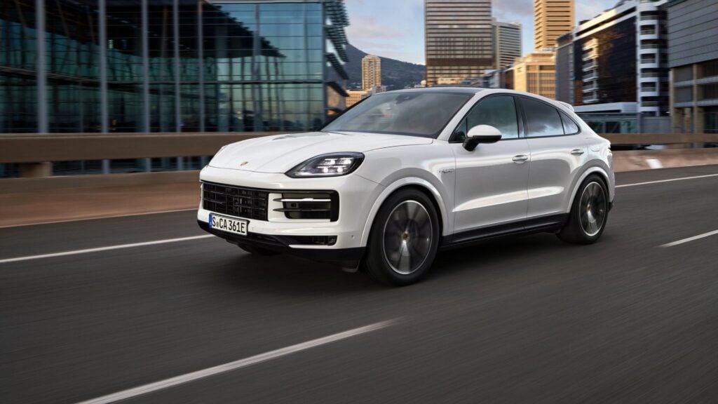 En vit Porsche Cayenne kör på en motorväg, omgiven av moderna byggnader i bakgrunden.