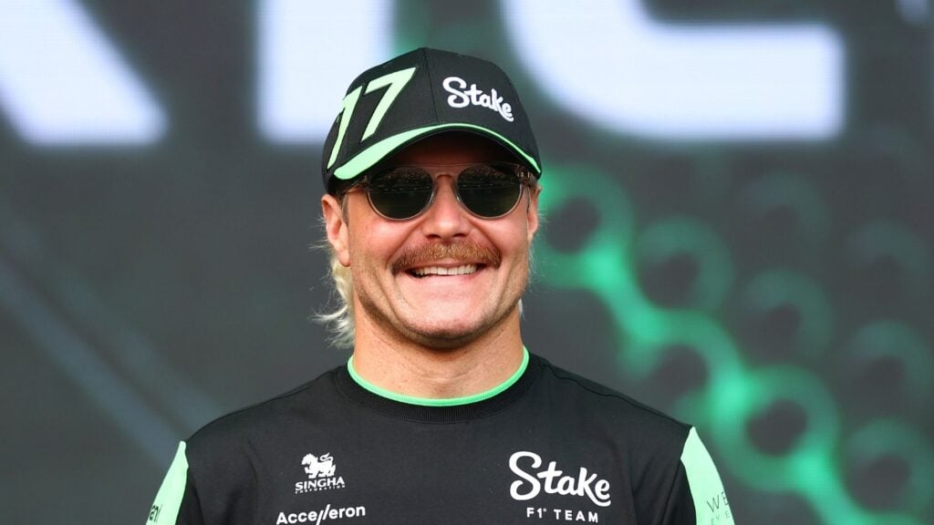 Valtteri Bottas ler stort i sin teamkeps och solglasögon under ett event, iförd sin racingoverall från Stake F1 Team.