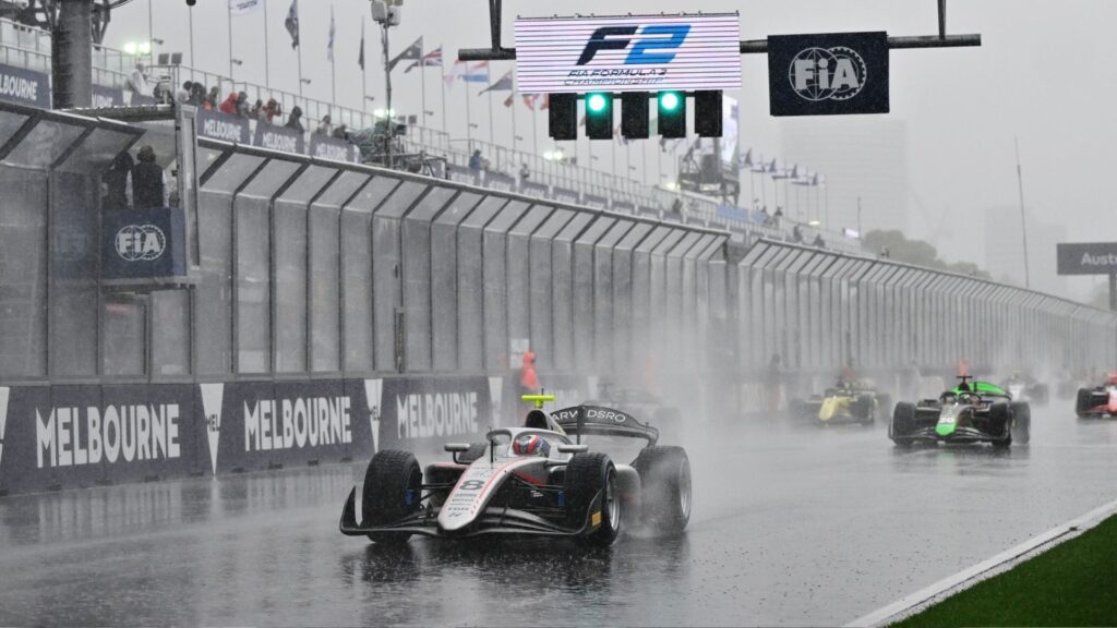 F2-bilar kör genom ösregnet på Albert Park Circuit i Melbourne, med kraftiga vattenstrålar bakom sig. Skyltarna för FIA och Formel 2 syns tydligt ovanför banan medan förarna försöker navigera i de svåra förhållandena.