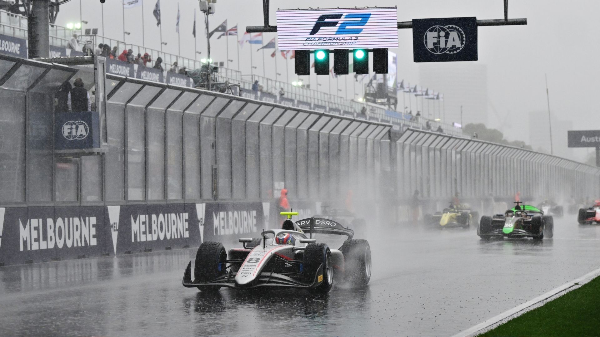 F2-bilar kör genom ösregnet på Albert Park Circuit i Melbourne, med kraftiga vattenstrålar bakom sig. Skyltarna för FIA och Formel 2 syns tydligt ovanför banan medan förarna försöker navigera i de svåra förhållandena.