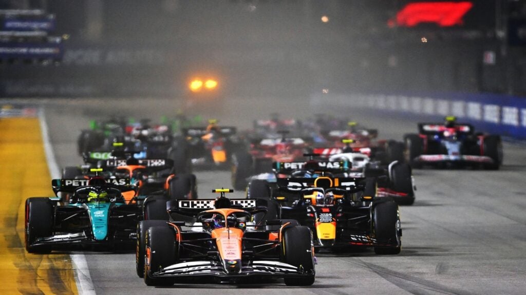 Formel 1-bilar i full fart genom en nattlig stadsbana, med en McLaren i ledningen tätt följd av en Red Bull, Mercedes och flera andra team. Strålkastarljus och glödande bromsar förstärker den intensiva racingstämningen.