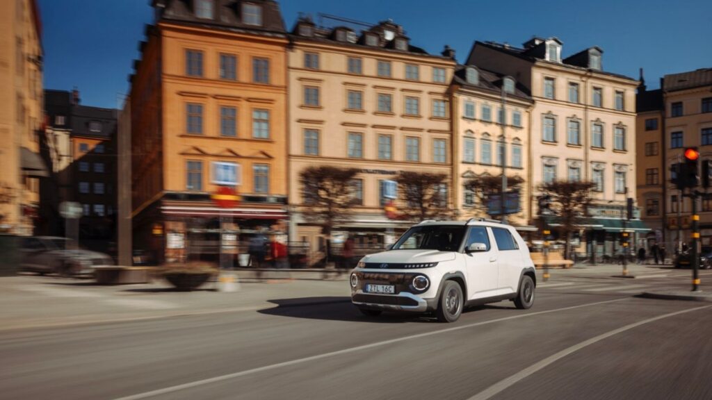 Vit Hyundai Inster kör genom Stockholm med stadshusfasader i bakgrunden.