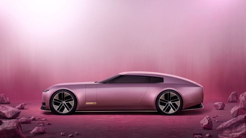 Sidoprofil av Jaguar Type 00 Concept, en futuristisk grand tourer i rosa metallic med aerodynamisk design och täckta hjul, omgiven av en ökenliknande miljö.