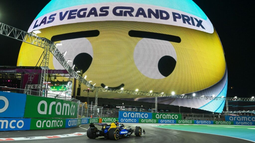 En Formel 1-bil kör genom en kurva på Las Vegas GP-banan, med den stora LED-kupolen Sphere i bakgrunden som visar en emoji.