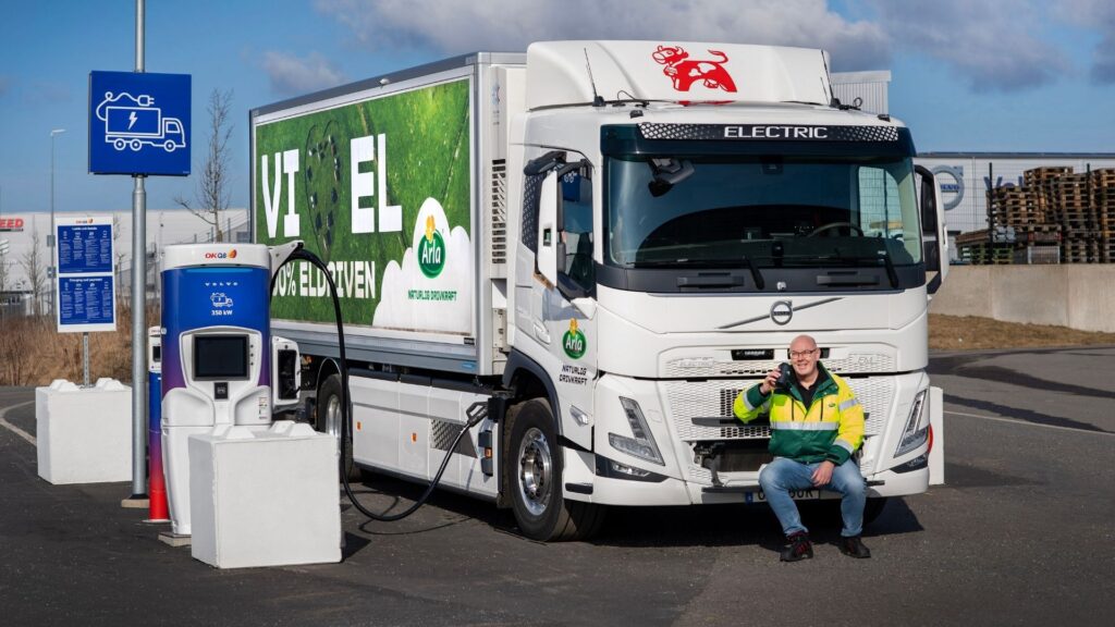 El-lastbil från Arla laddas vid en Volvo- och OKQ8-laddstation. Chaufför i gulgrön jacka sitter framför lastbilen och håller upp sin mobiltelefon.