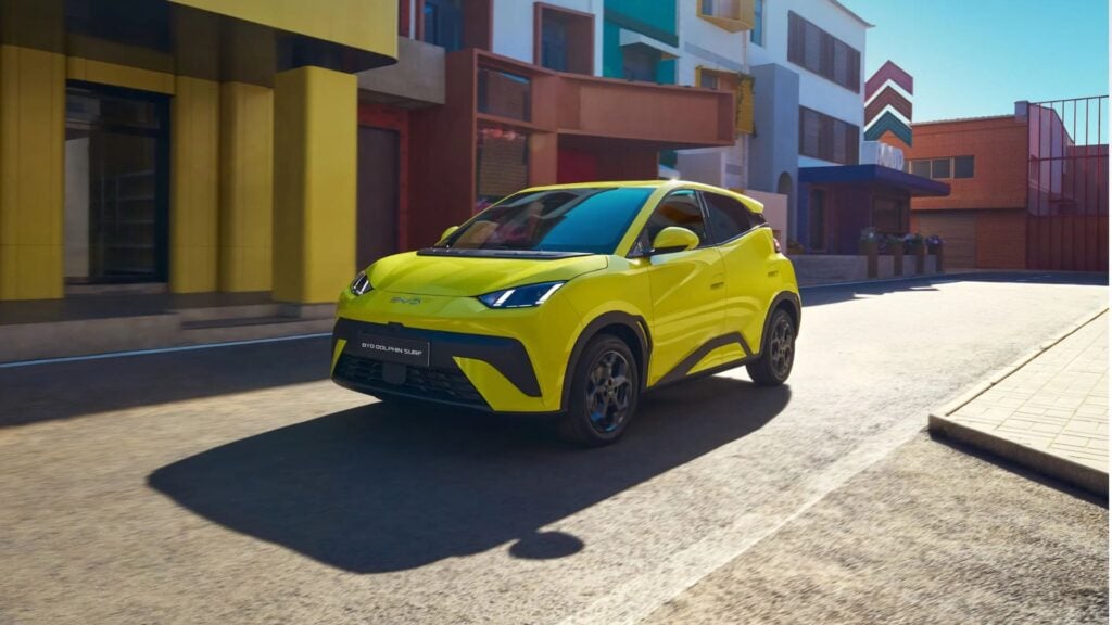 BYD Dolphin Surf kör längs färgglad stadsmiljö i dagsljus, gul bil sedd snett framifrån.