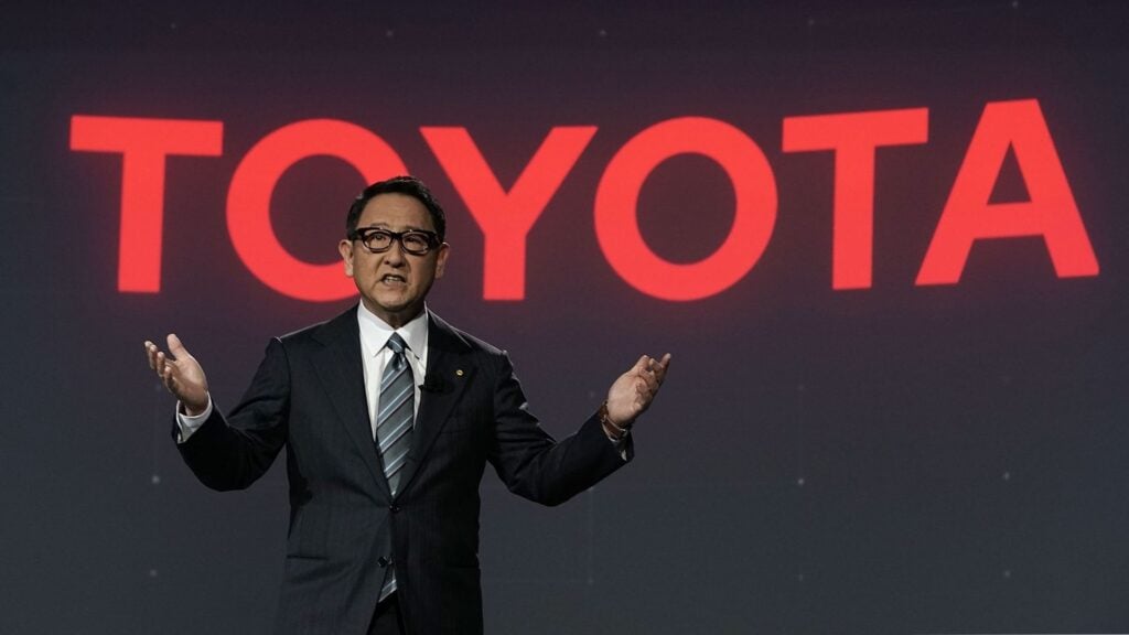 Akio Toyoda står framför en stor Toyota-logga under ett presentationstillfälle.