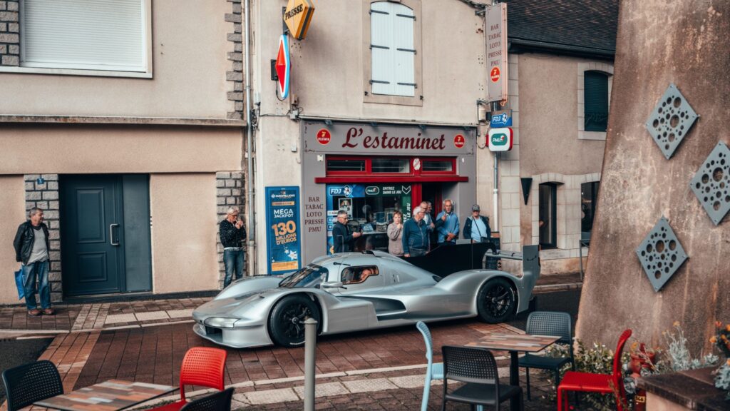 Porsche 963 RSP parkerad framför baren L’Estaminet i centrala Le Mans, omgiven av nyfikna förbipasserande.