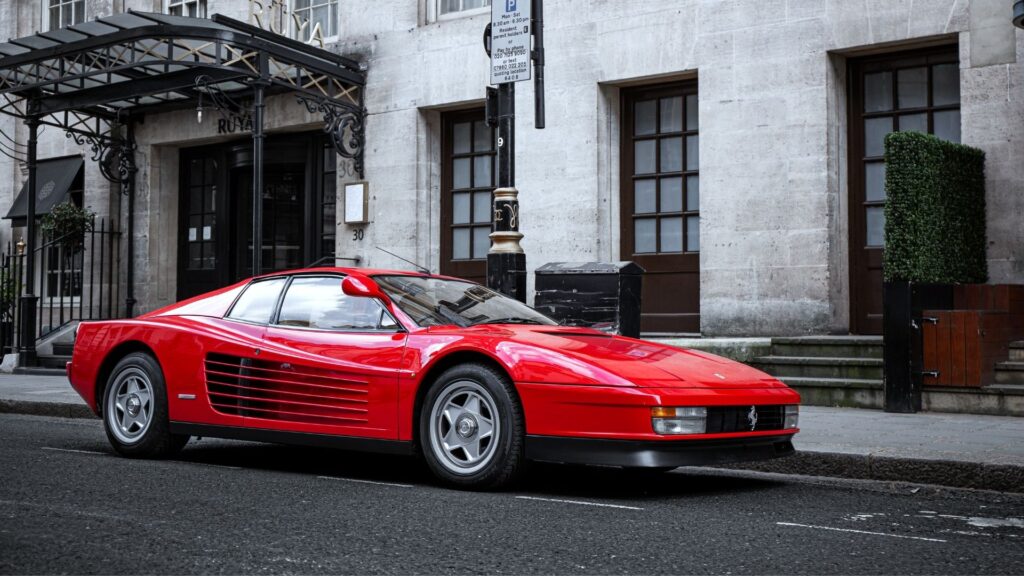 Röd Ferrari Testarossa parkerad på stadsgata framför stenfasad.