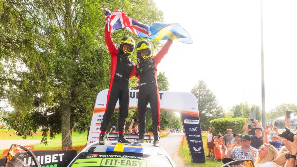 Oliver Solberg och Elliott Edmondson står på sin Toyota och firar WRC-seger med svenska och brittiska flaggan