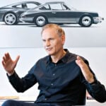 Oliver Blume gestikulerar i en intervju sittandes framför Porsche-konst på väggen.