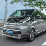 Grå Kia PV5 snett framifrån körandes på en strandväg.