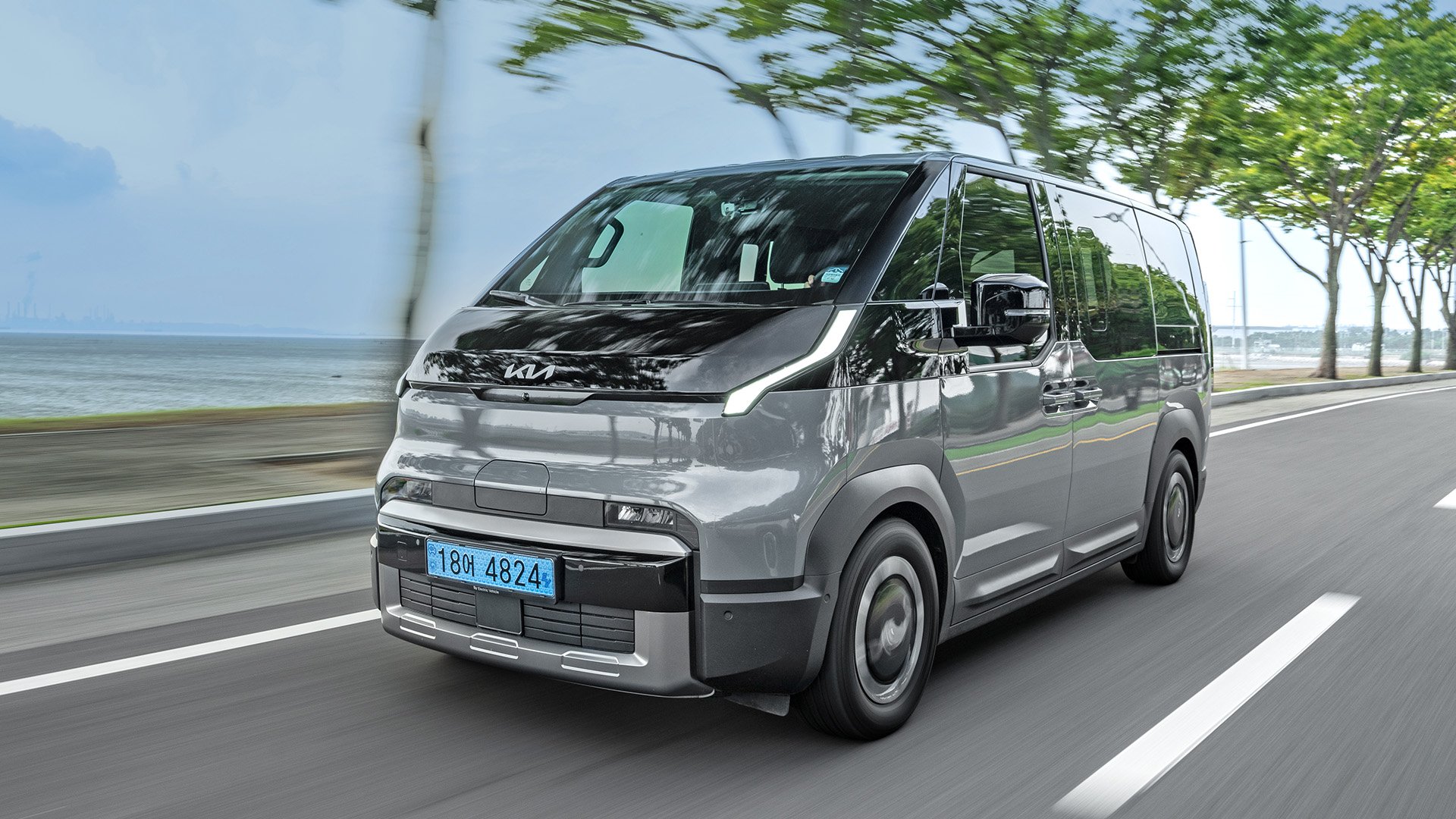 Grå Kia PV5 snett framifrån körandes på en strandväg.