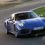 Blå Porsche Turbo S snett framifrån på motorbana med solnedgång i bakgrunden.