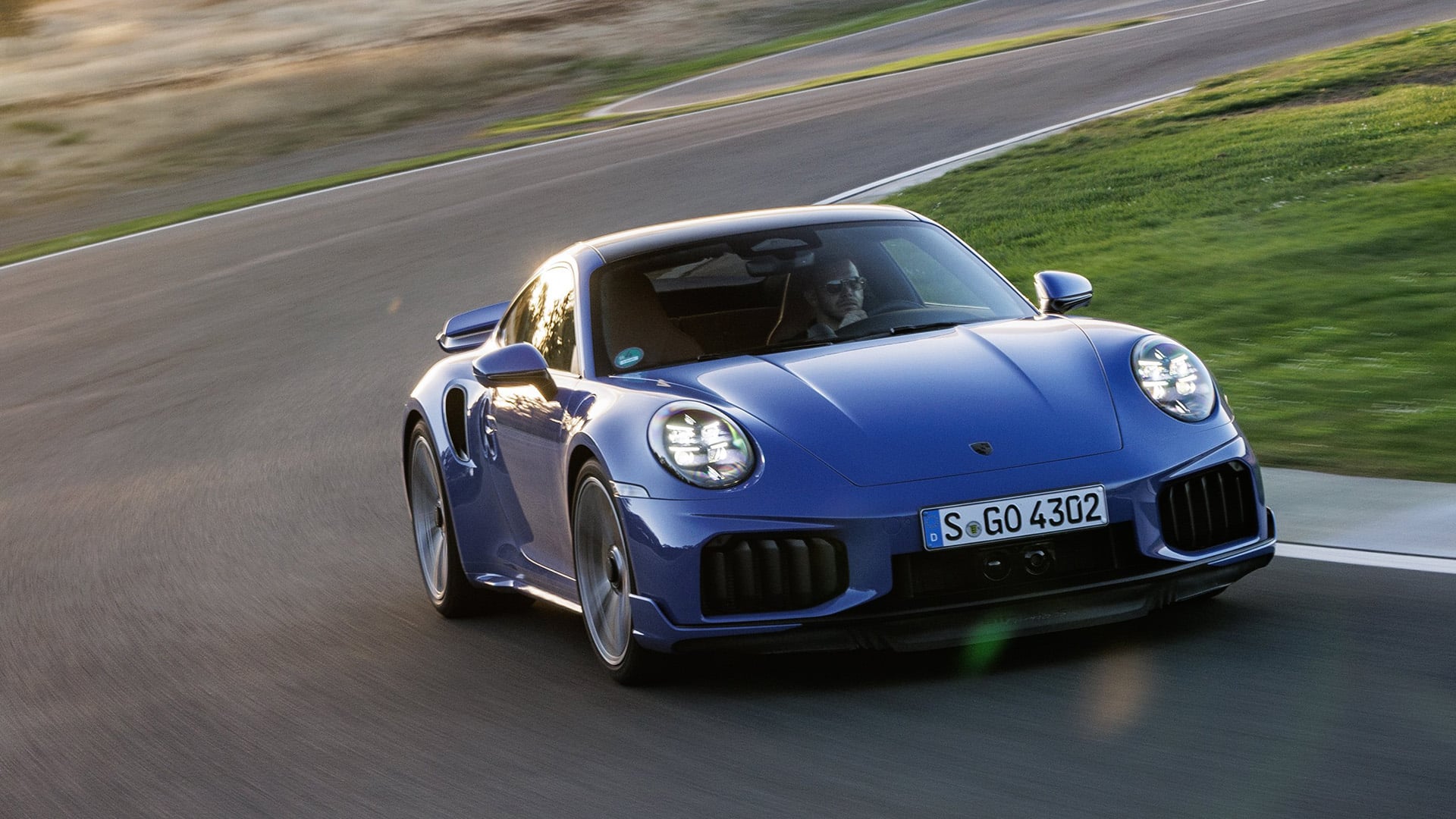 Blå Porsche Turbo S snett framifrån på motorbana med solnedgång i bakgrunden.
