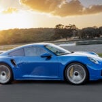 Blå Porsche Turbo S i profil på motorbana med solnedgång i bakgrunden.