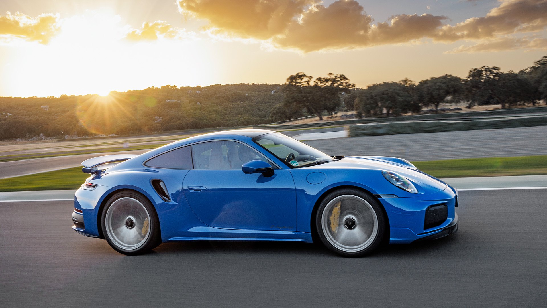 Blå Porsche Turbo S i profil på motorbana med solnedgång i bakgrunden.