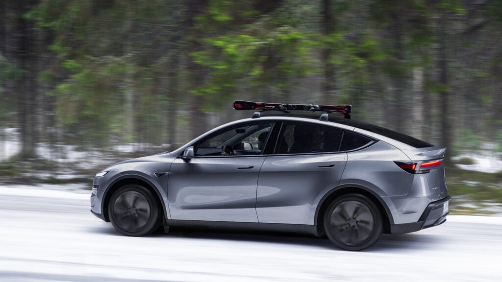 Grå Tesla Model Y på snöig skogsväg i profil. Med takräcke och skidor på taket.
