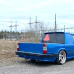 Blå Volvo 740 A-traktor snett bakifrån på grusväg med elledningar i bakgrunden.