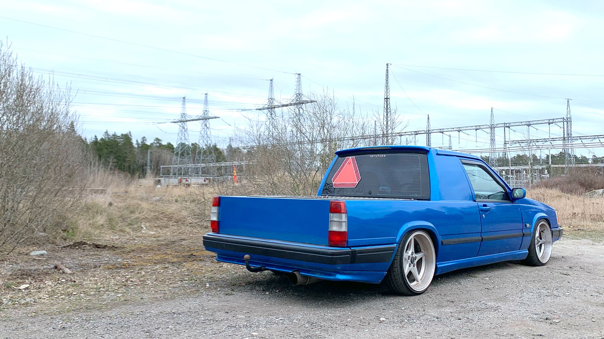 Blå Volvo 740 A-traktor snett bakifrån på grusväg med elledningar i bakgrunden.
