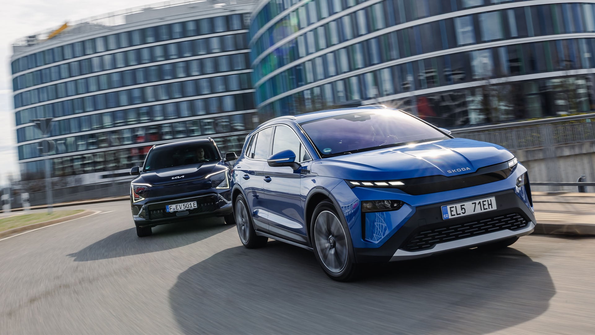 Test Kia EV5 vs Skoda Elroq. Skoda längst fram i bild och i bakgrunden är stora byggnader täckta med glasfönster.