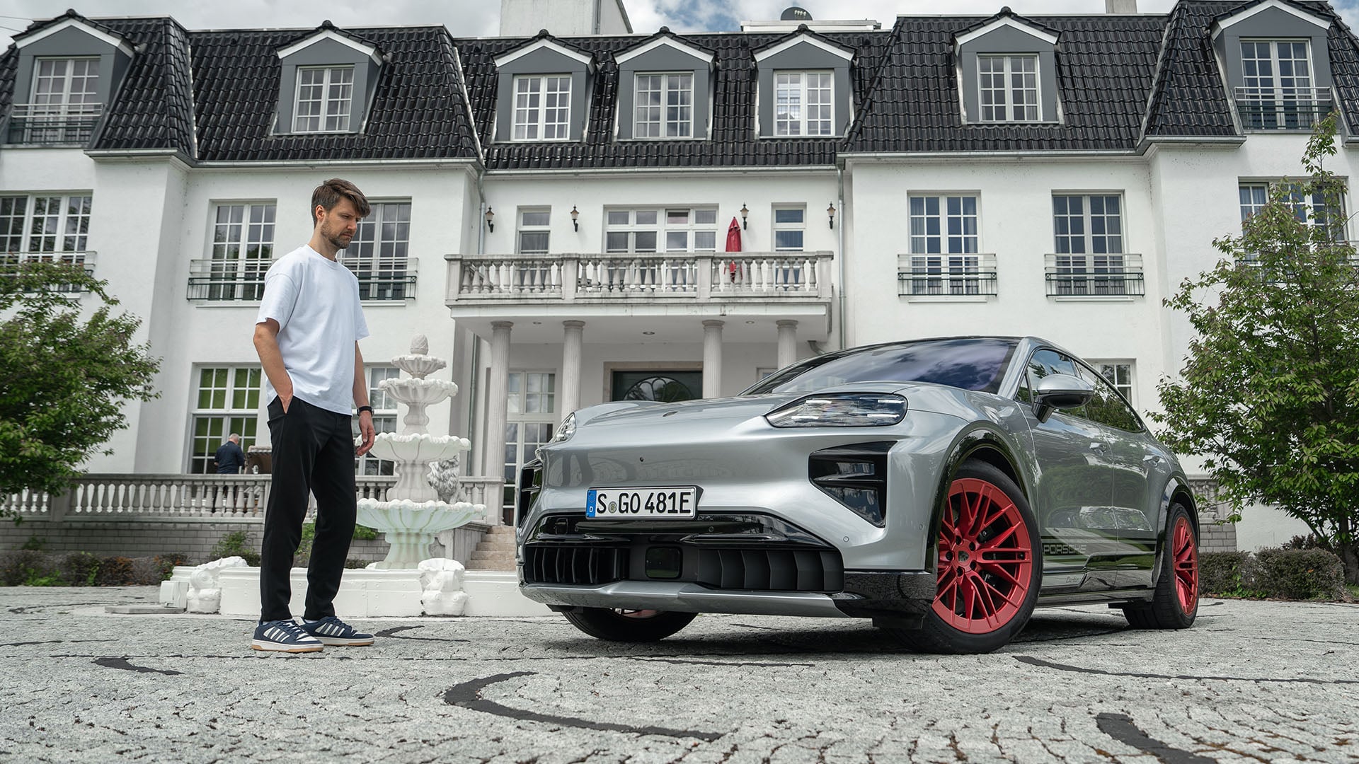 John Sempill till vänster står och tittar på en silver Porsche Cayenne Coupé Electric som står framför ett hus.