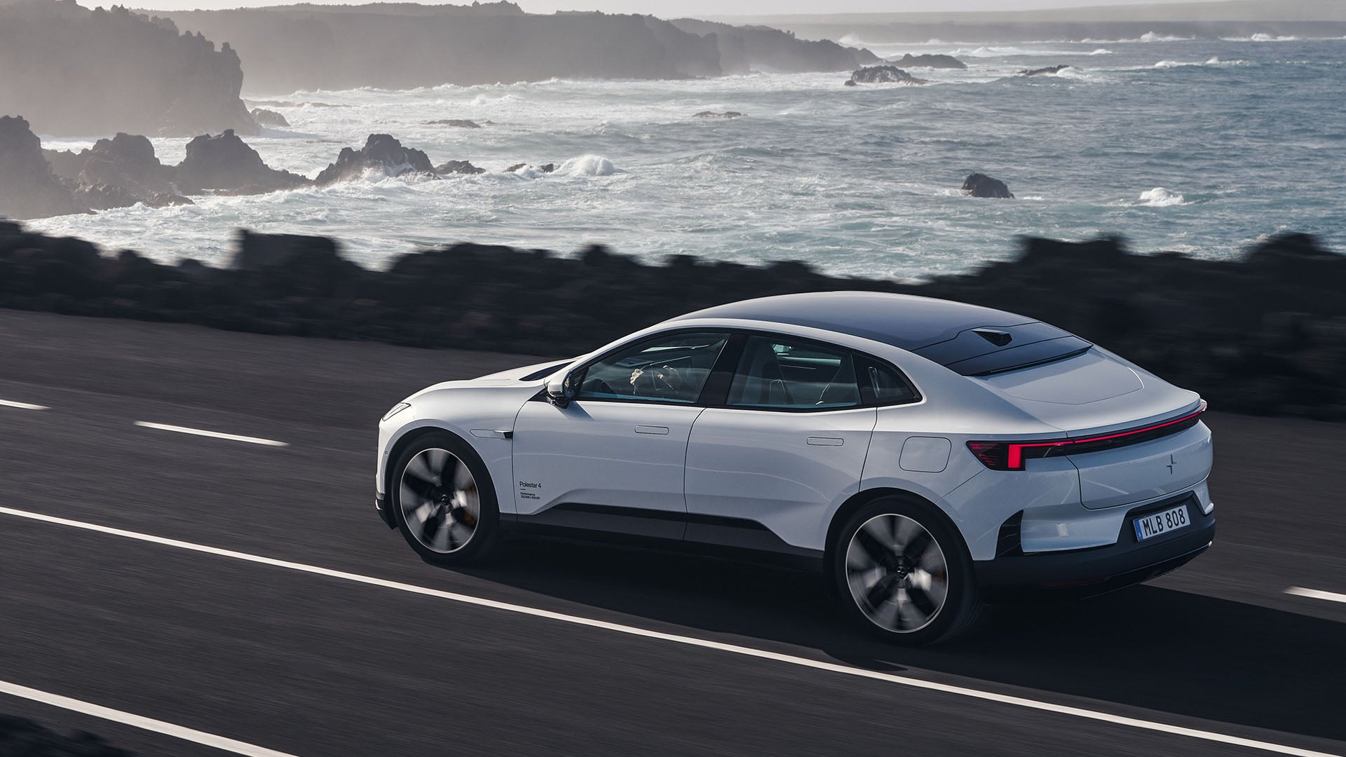 Vit Polestar 4 på snett bakifrån på strandväg med hav i bakgrunden.