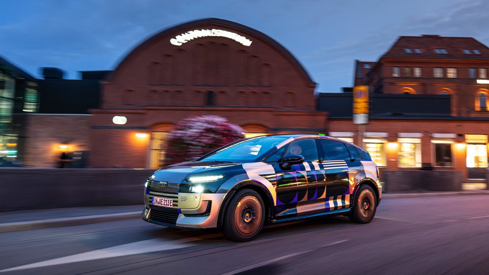 Lätt kamouflerad Audi A2 e-tron snett framifrån körandes på väg i stan.