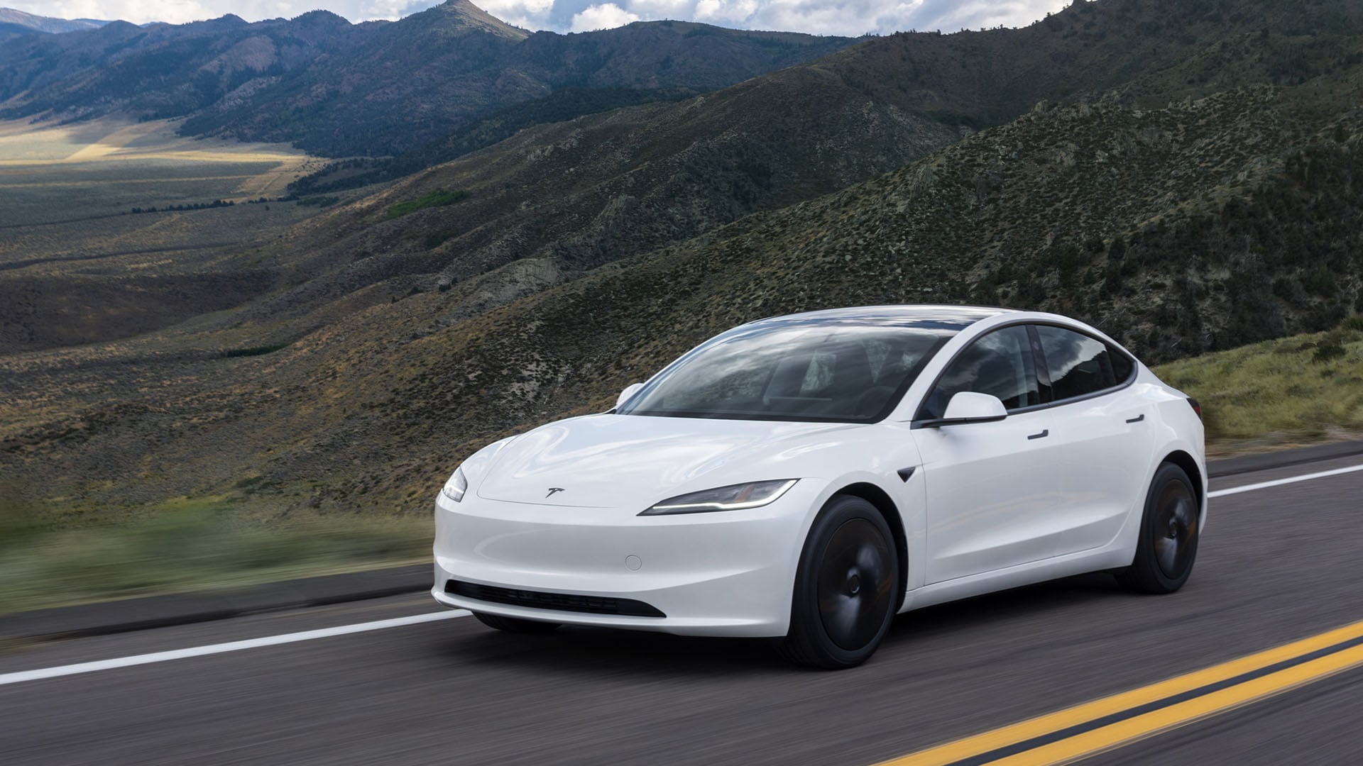 Vit Tesla Model 3 snett framifrån på väg med landskap i bakgrunden.