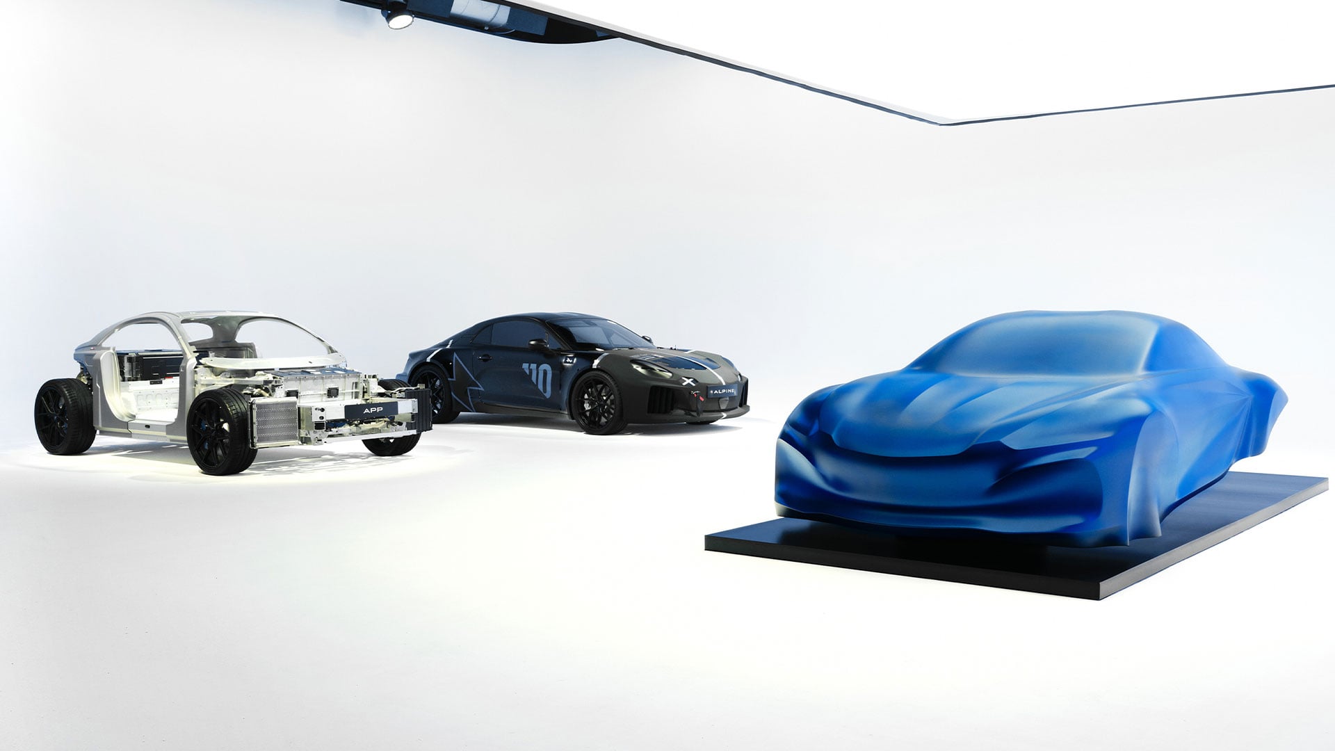 Blå skulptur av kommande Alpine A110 i studiomiljö, med ett testchassi och en maskerad testbil i bakgrunden.