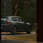 Grön Volvo EX60 syns genom ett fönster med skog i bakgrunden.