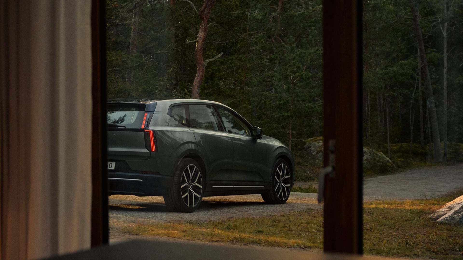 Grön Volvo EX60 syns genom ett fönster med skog i bakgrunden.