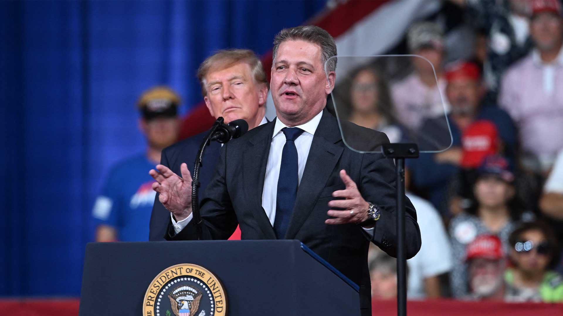 Audi-chefen Gernot Döllner talar vid en talarstol på Donald Trumps valmöte i Georgia, med Trump stående bakom honom.