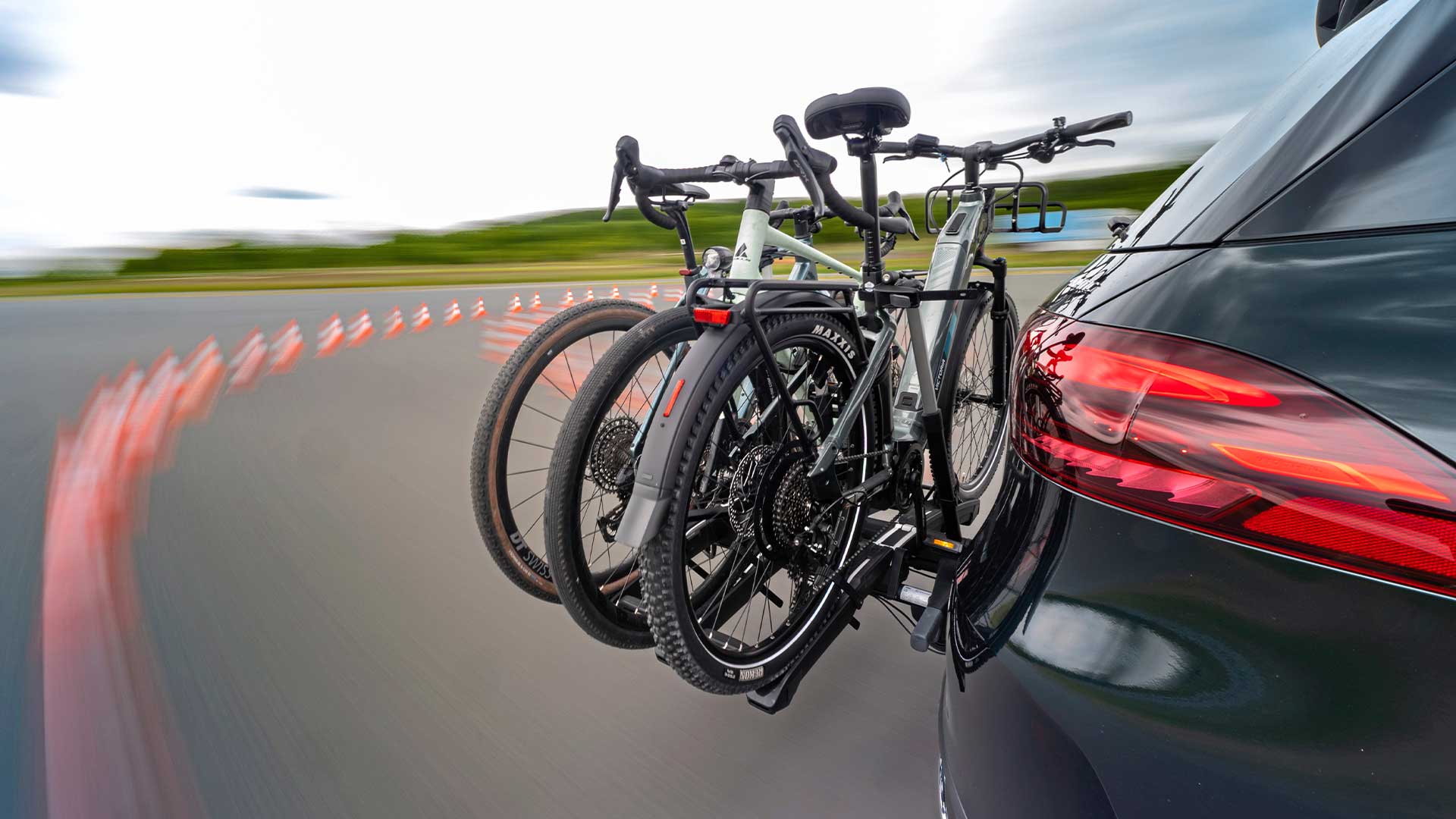 Tre cyklar på en dragkroksmonterad cykelhållare bakom en bil under körning på en bana med koner.