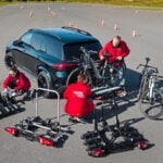 Tre personer i röda jackor monterar och lastar cyklar på flera dragkroksmonterade cykelhållare bakom en Mercedes GLE på en testbana med koner.