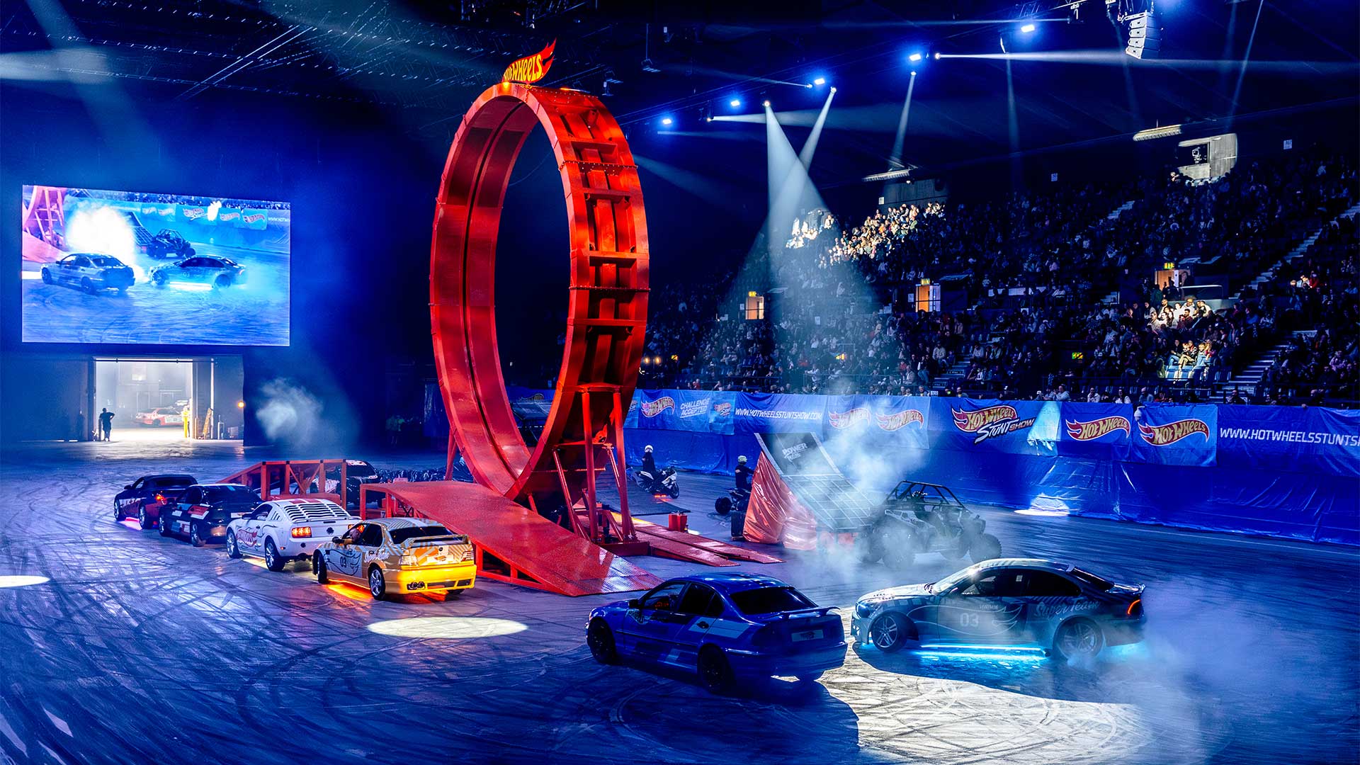 Hot Wheels-stuntshow i arena med flera bilar, rök, publik och en stor loop på banan.