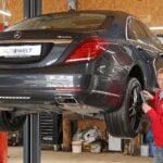 Mörkgrå Mercedes-Benz S-Klass W222 inspekteras av man i röd jacka medan den hänger på en billyft en i verkstad.