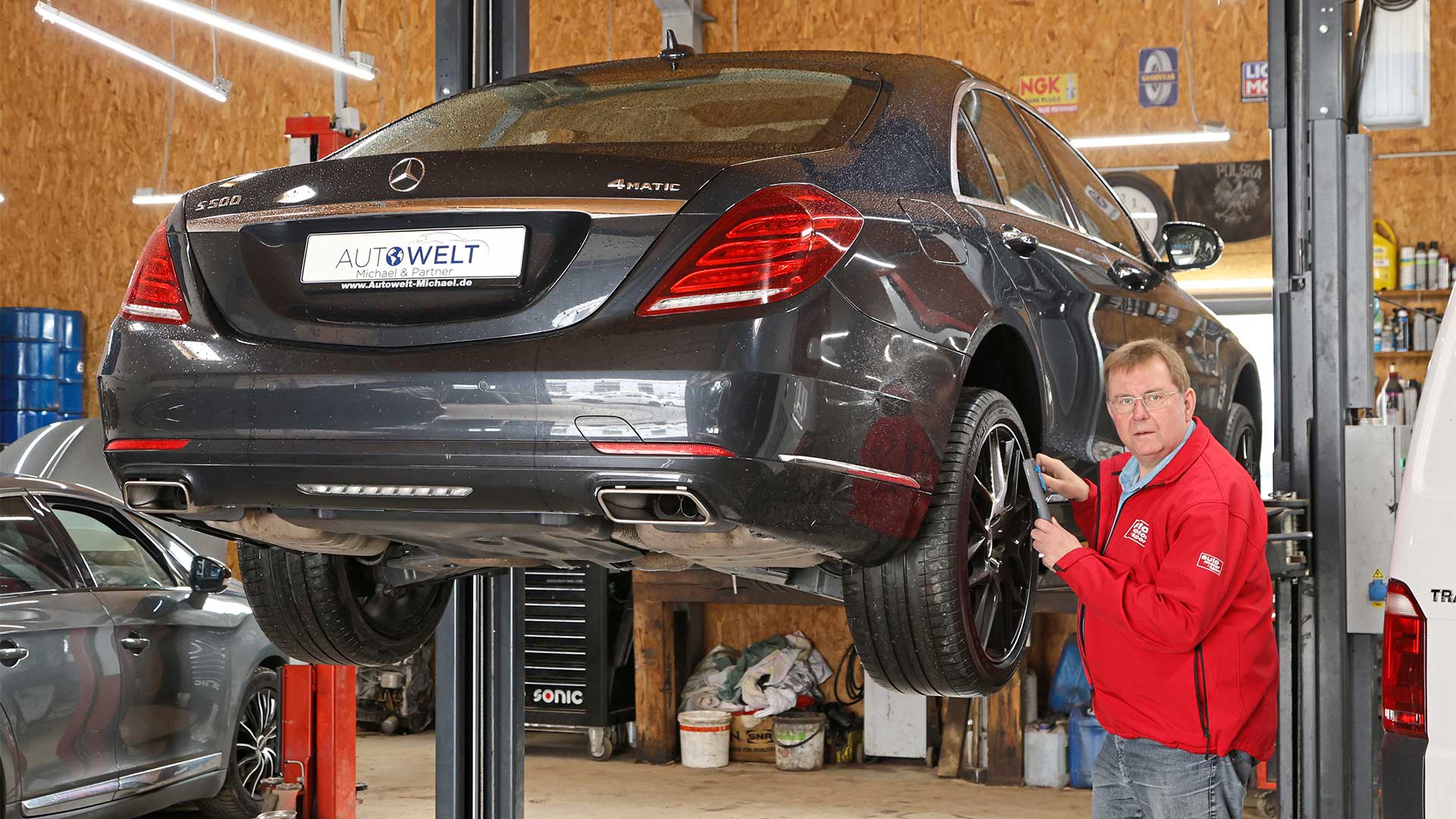 Mörkgrå Mercedes-Benz S-Klass W222 inspekteras av man i röd jacka medan den hänger på en billyft en i verkstad.